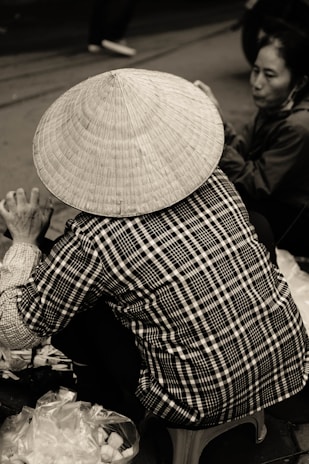 A person wearing a traditional conical hat is seated on a stool, facing away from the camera. The individual is dressed in a checkered shirt and is engaged in an activity, possibly involving plastic bags and items on the ground. Another person is partially visible in the background, sitting on the ground, attentively involved in the same activity.