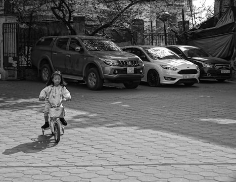 A young child wearing a headband and casual clothes is riding a small bicycle with training wheels on a paved area. In the background, there are several parked cars and a fenced area with trees.