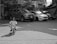 A young child wearing a headband and casual clothes is riding a small bicycle with training wheels on a paved area. In the background, there are several parked cars and a fenced area with trees.
