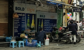 A small Vietnamese street food stall is set up next to a wall covered with large advertisements for Hanoi Bold and Hanoi Light beers. Several low plastic stools are placed around, and a vendor sits nearby. Various items and bags are scattered on the ground. Motorbikes are parked on the street, and a group of people gather in the background near colorful balloons.