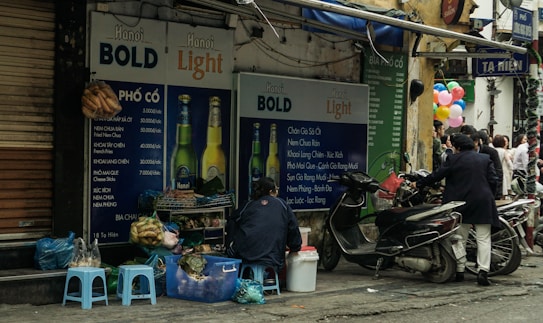 A small Vietnamese street food stall is set up next to a wall covered with large advertisements for Hanoi Bold and Hanoi Light beers. Several low plastic stools are placed around, and a vendor sits nearby. Various items and bags are scattered on the ground. Motorbikes are parked on the street, and a group of people gather in the background near colorful balloons.