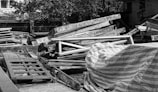 A pile of wooden planks and frames is stacked in an outdoor setting, some covered with a striped tarp. There are trees and buildings in the background.