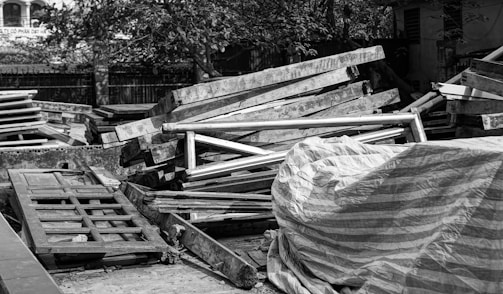 A pile of wooden planks and frames is stacked in an outdoor setting, some covered with a striped tarp. There are trees and buildings in the background.