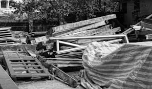 A pile of wooden planks and frames is stacked in an outdoor setting, some covered with a striped tarp. There are trees and buildings in the background.