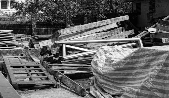 A pile of wooden planks and frames is stacked in an outdoor setting, some covered with a striped tarp. There are trees and buildings in the background.