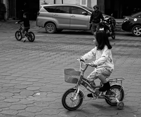 A young girl rides a small bicycle with training wheels on a paved area. She is dressed in light-colored, patterned clothing. In the background, a child rides another bicycle, and an adult stands near a motorcycle and cars are parked nearby.