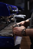 A close-up view of a person using an espresso machine to prepare a beverage. The individual is holding a metal steaming pitcher under the steam wand, and the espresso machine is prominently blue with shiny metallic parts.