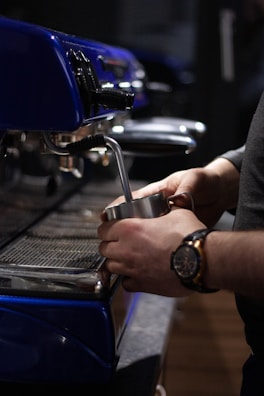 A close-up view of a person using an espresso machine to prepare a beverage. The individual is holding a metal steaming pitcher under the steam wand, and the espresso machine is prominently blue with shiny metallic parts.