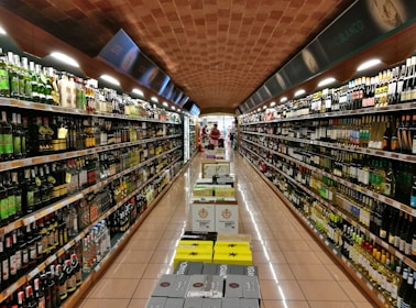 A supermarket aisle lined with shelves filled with a diverse selection of bottles and boxes, primarily alcoholic beverages. The ceiling is arched with a brick pattern and the floor is polished tile. At the far end of the aisle, there are people shopping.