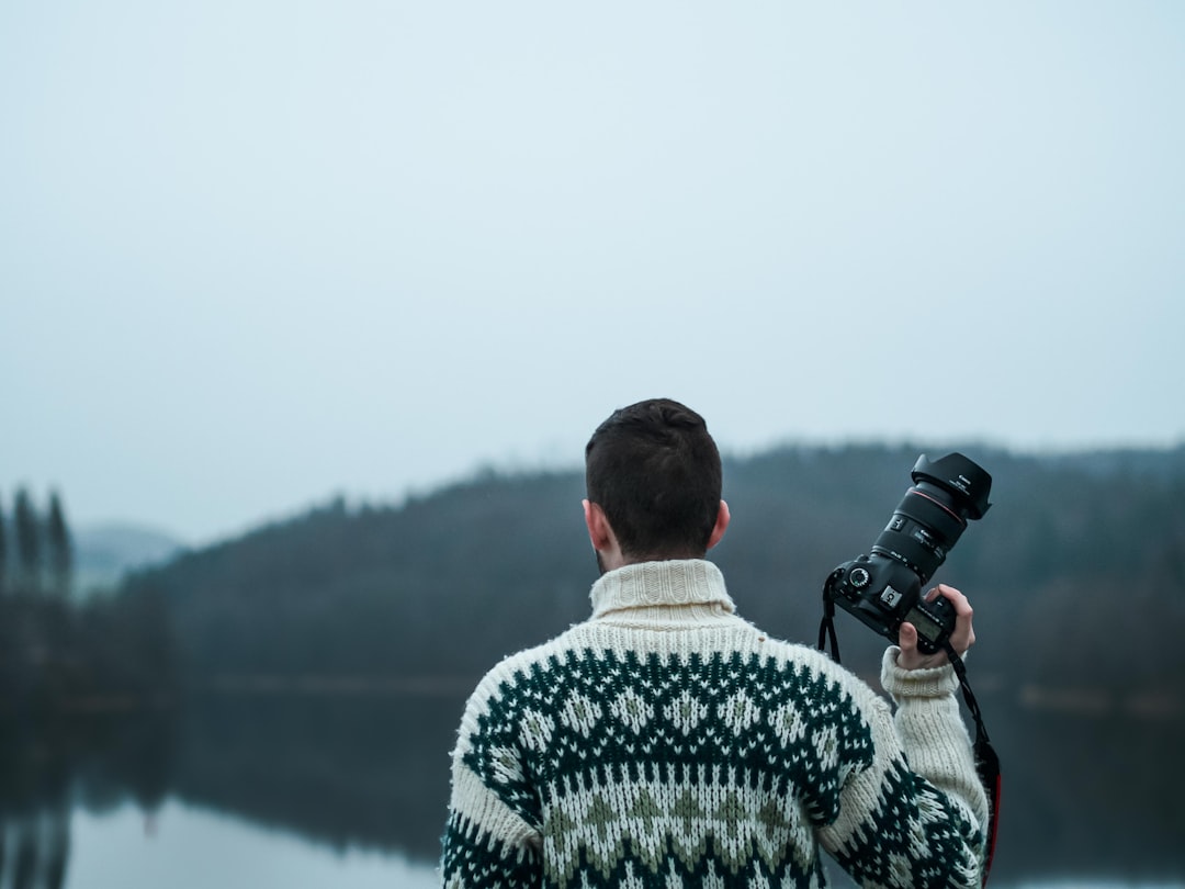 man carrying a black DSLR camera facing a body of water