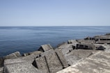 A calm, expansive ocean stretches out to the horizon under a clear blue sky. Foreground features large, geometric concrete blocks forming a breakwater along the shore.