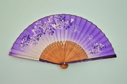 A decorative hand fan with a curved shape and an intricate design. The fan displays a gradient from white to purple with floral patterns and butterflies. The handle and ribs are made of a light brown wood with detailed carvings.