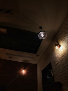 A dimly lit room with brick and white textured walls, featuring three light fixtures. One ceiling light with a wire cage design and two wall-mounted lights create a warm ambiance. The lighting casts soft shadows on the walls.