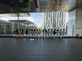 A group of children in colorful outfits stand in a line facing a large glass window, looking out at a cityscape with modern buildings. The ceiling reflects the scenery and the tiled floor adds a geometric pattern to the scene.