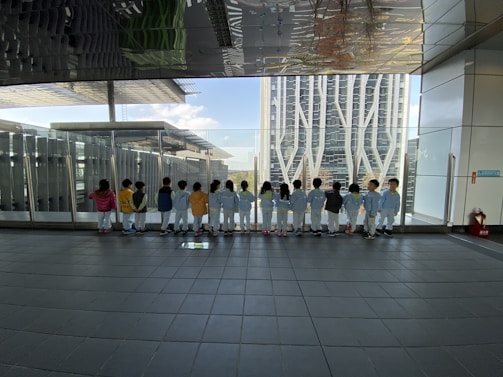 A group of children in colorful outfits stand in a line facing a large glass window, looking out at a cityscape with modern buildings. The ceiling reflects the scenery and the tiled floor adds a geometric pattern to the scene.