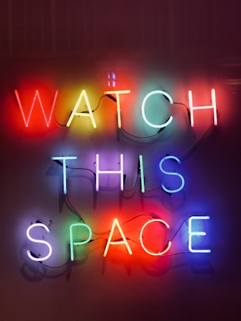 Neon lights form the words 'WATCH THIS SPACE' with each letter in vibrant, bold colors against a dark background. The glow of the neon creates a lively and eye-catching effect.