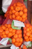 Several mesh bags filled with bright orange tangerines are stacked together. The tangerines are visible through the mesh, which gives a textured appearance to the fruit. Each bag has a label attached, adding a commercial aspect to the image.