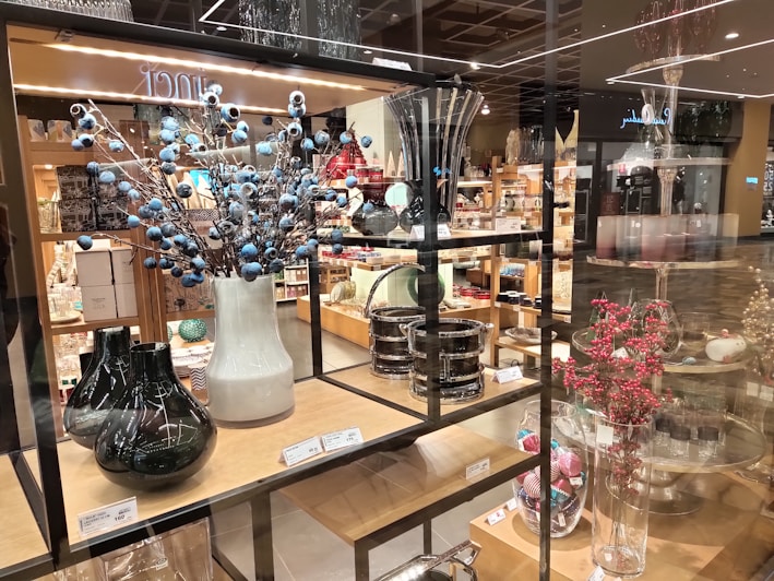 A store display featuring a variety of decorative vases and glassware arranged on wooden shelves. There are tall vases with blue and red berry-like adornments and assorted glass containers. The shelves are stacked with neatly arranged home decor items with price tags visible.
