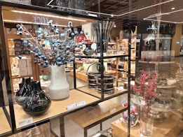 A store display featuring a variety of decorative vases and glassware arranged on wooden shelves. There are tall vases with blue and red berry-like adornments and assorted glass containers. The shelves are stacked with neatly arranged home decor items with price tags visible.