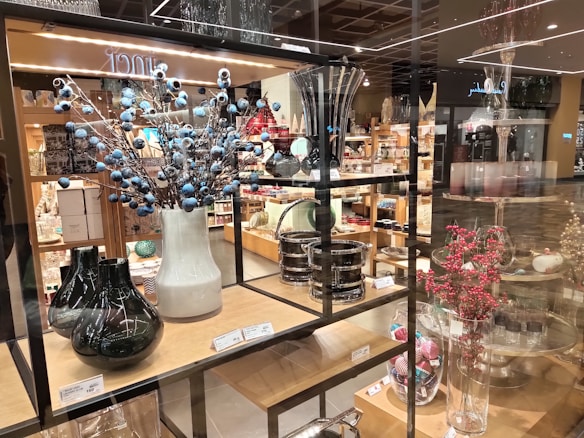 A store display featuring a variety of decorative vases and glassware arranged on wooden shelves. There are tall vases with blue and red berry-like adornments and assorted glass containers. The shelves are stacked with neatly arranged home decor items with price tags visible.