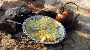A rustic outdoor cooking setup featuring a metal plate filled with scrambled eggs resting on a bed of coals surrounded by rocks. A well-used, blackened kettle is positioned next to the plate. There is evidence of cracked eggshells nearby, and the scene is set on a rough, earthy surface.
