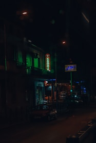 A dimly lit street scene during nighttime featuring a neon pharmacy sign glowing in green. The street is lined with a few parked cars and a building with lighted windows in the background. Streetlights create a moody ambiance, reflecting off the damp road surface.
