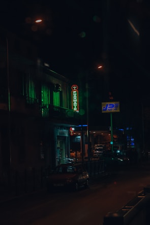 A dimly lit street scene during nighttime featuring a neon pharmacy sign glowing in green. The street is lined with a few parked cars and a building with lighted windows in the background. Streetlights create a moody ambiance, reflecting off the damp road surface.