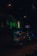 A dimly lit street scene during nighttime featuring a neon pharmacy sign glowing in green. The street is lined with a few parked cars and a building with lighted windows in the background. Streetlights create a moody ambiance, reflecting off the damp road surface.