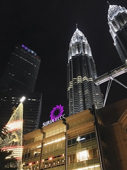 Illuminated skyscrapers dominate the night sky, with the striking twin towers prominently featured. Below, a well-lit shopping center entrance displays the sign 'SURIA KLCC'. A festive Christmas tree adorned with glowing lights adds a holiday touch to the urban scene.