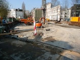 A construction site is visible with an excavator and other construction machinery. There are construction barriers and temporary structures, with the partially demolished facade of a building supported by scaffolding. Trees and buildings surround the site under a clear blue sky.
