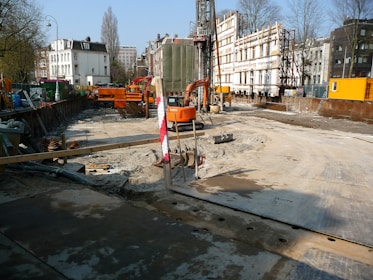 A construction site is visible with an excavator and other construction machinery. There are construction barriers and temporary structures, with the partially demolished facade of a building supported by scaffolding. Trees and buildings surround the site under a clear blue sky.