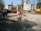 A construction site is visible with an excavator and other construction machinery. There are construction barriers and temporary structures, with the partially demolished facade of a building supported by scaffolding. Trees and buildings surround the site under a clear blue sky.