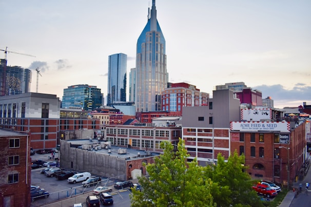 The image captures a cityscape with modern skyscrapers and historic brick buildings. A prominent blue and glass tower stands in the center, surrounded by other tall buildings. In the foreground, there is a parking lot and a building with a sign reading 'ACME FEED & SEED'. The scene includes some greenery from trees and a few cars parked on the street.