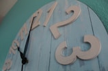 A large wall clock has a rustic design with wooden planks painted in light blue. The clock's numbers are raised and painted in a cream color. The hour and minute hands are black with a simple design, positioned between 9 and 10. The background wall is painted in teal.