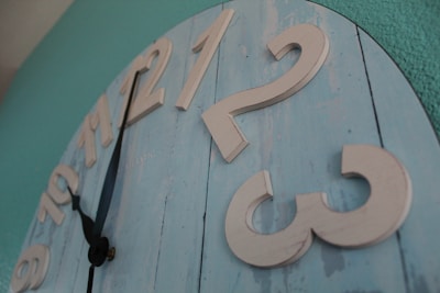 A large wall clock has a rustic design with wooden planks painted in light blue. The clock's numbers are raised and painted in a cream color. The hour and minute hands are black with a simple design, positioned between 9 and 10. The background wall is painted in teal.
