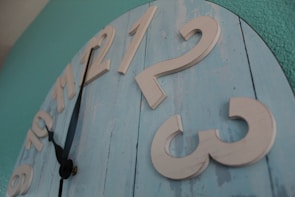A large wall clock has a rustic design with wooden planks painted in light blue. The clock's numbers are raised and painted in a cream color. The hour and minute hands are black with a simple design, positioned between 9 and 10. The background wall is painted in teal.