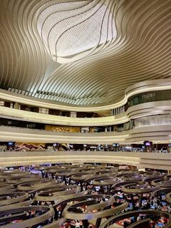 An architectural space with a large, intricately designed ceiling featuring wavy patterns. Multiple floors with balconies overlook a bustling gaming or trading floor with numerous people and electronic displays.