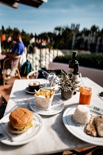 A table is set with a variety of food items including a hamburger, fries in a metal basket, a plate of rice with grilled chicken, a cappuccino, and an orange drink garnished with a slice of citrus. There is a small potted plant on the table for decoration. In the background, a person is sitting at a table, and there are several flags visible along with a garden area.