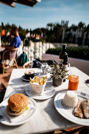 A table is set with a variety of food items including a hamburger, fries in a metal basket, a plate of rice with grilled chicken, a cappuccino, and an orange drink garnished with a slice of citrus. There is a small potted plant on the table for decoration. In the background, a person is sitting at a table, and there are several flags visible along with a garden area.