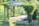 A wooden pergola structure covered with lush green vines and surrounded by a well-maintained garden. There are smooth tree stump seats and a small path leading to a flower bed with mixed flowers, all set against a backdrop of bright green grass.