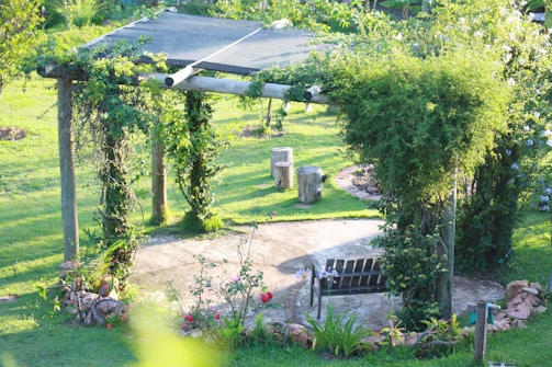 A wooden pergola structure covered with lush green vines and surrounded by a well-maintained garden. There are smooth tree stump seats and a small path leading to a flower bed with mixed flowers, all set against a backdrop of bright green grass.