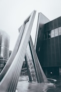 A modern architectural structure featuring tall, angled metal beams that form an arch, situated in an urban setting. The background shows part of a building with large windows and a cloudy, overcast sky. The scene has a monochromatic tone, emphasizing the sleek lines and geometric shapes of the structure.