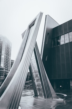 A modern architectural structure featuring tall, angled metal beams that form an arch, situated in an urban setting. The background shows part of a building with large windows and a cloudy, overcast sky. The scene has a monochromatic tone, emphasizing the sleek lines and geometric shapes of the structure.