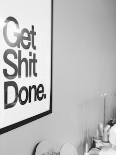 A black and white poster with bold text reading 'Get Shit Done' is mounted on a wall. To the right, blurry details of a workspace can be seen, including several items such as bottles and rolls, all slightly out of focus.