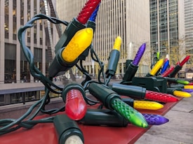 Giant, colorful Christmas lights are arranged in an outdoor urban setting, with large buildings in the background. The oversized bulbs are of various colors including red, yellow, green, purple, and blue, and are connected by thick, dark green cables.