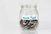 A clear glass jar filled with various coins, labeled with a handwritten note that reads 'House Fund.' The jar is simple in design, placed against a plain white background.
