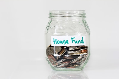 A clear glass jar filled with various coins, labeled with a handwritten note that reads 'House Fund.' The jar is simple in design, placed against a plain white background.