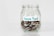 A clear glass jar filled with various coins, labeled with a handwritten note that reads 'House Fund.' The jar is simple in design, placed against a plain white background.