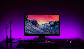 A desktop computer setup featuring a monitor displaying a scenic landscape with snow-covered mountains under a vibrant sunset sky. The setup is backlit with purple LED lighting, creating a striking contrast. A mechanical keyboard with colored backlighting and a gaming mouse are positioned on a wooden desk. Minimalist lamps and other small items are placed to the left of the monitor.