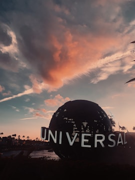A globe structure with the word 'Universal' is seen against the backdrop of a picturesque sky filled with soft, colorful clouds during sunset. Silhouettes of palm trees line the horizon, adding to the tranquil and cinematic ambiance.